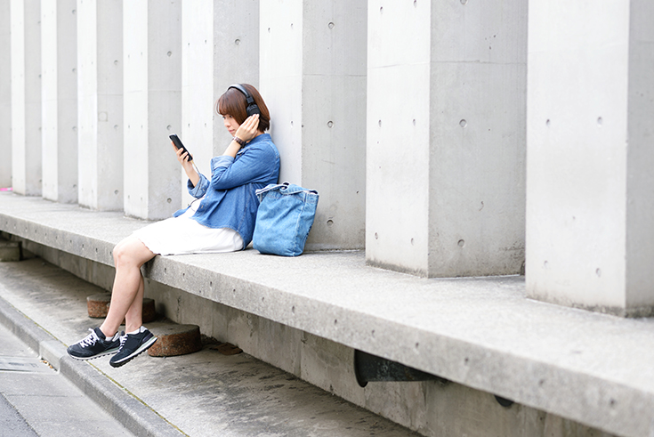 Photographie d'une jeune femme écoutant un fichier audio (radio, podcast) avec son casque tout en regardant son smartphone.