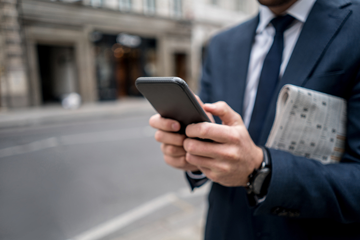 Photographie d'un homme en costume dans la rue; tenant à la main son smartphone et sous le bras son smartphone.