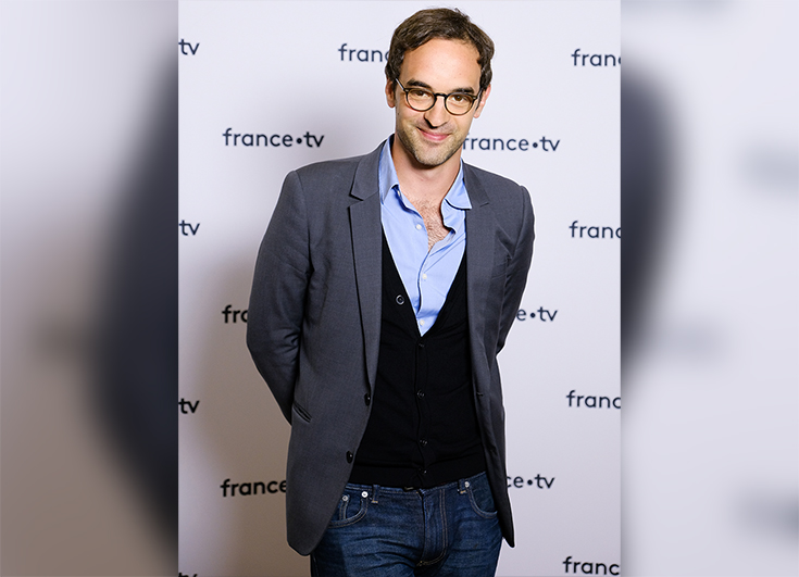 Portrait d'Antonio Grigolini, directeur éditorial de l'offre France.tv Slash. Il porte une chemise bleu clair ouverte, avec un cardigan bleu marine et une veste de costume anthracite, sur un jean. 